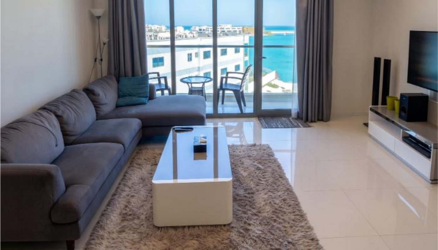 Modern living room with a gray sofa, white coffee table, rug, TV unit, and balcony doors overlooking a waterfront view on a sunny day.