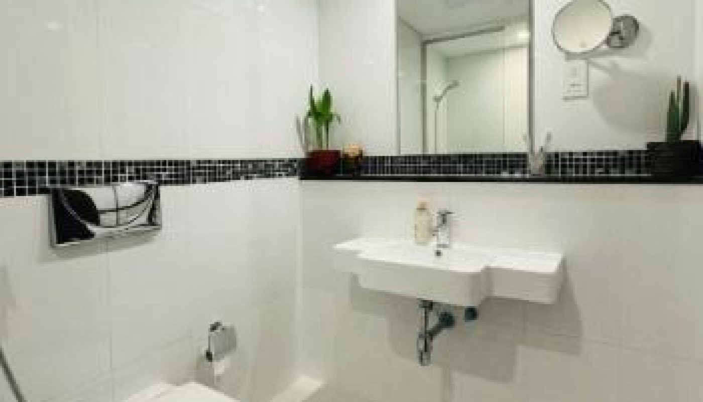 Modern bathroom with white tiles, wall-mounted toilet, sink with mirror above, small plants on a shelf, and a blue patterned rug on the floor.