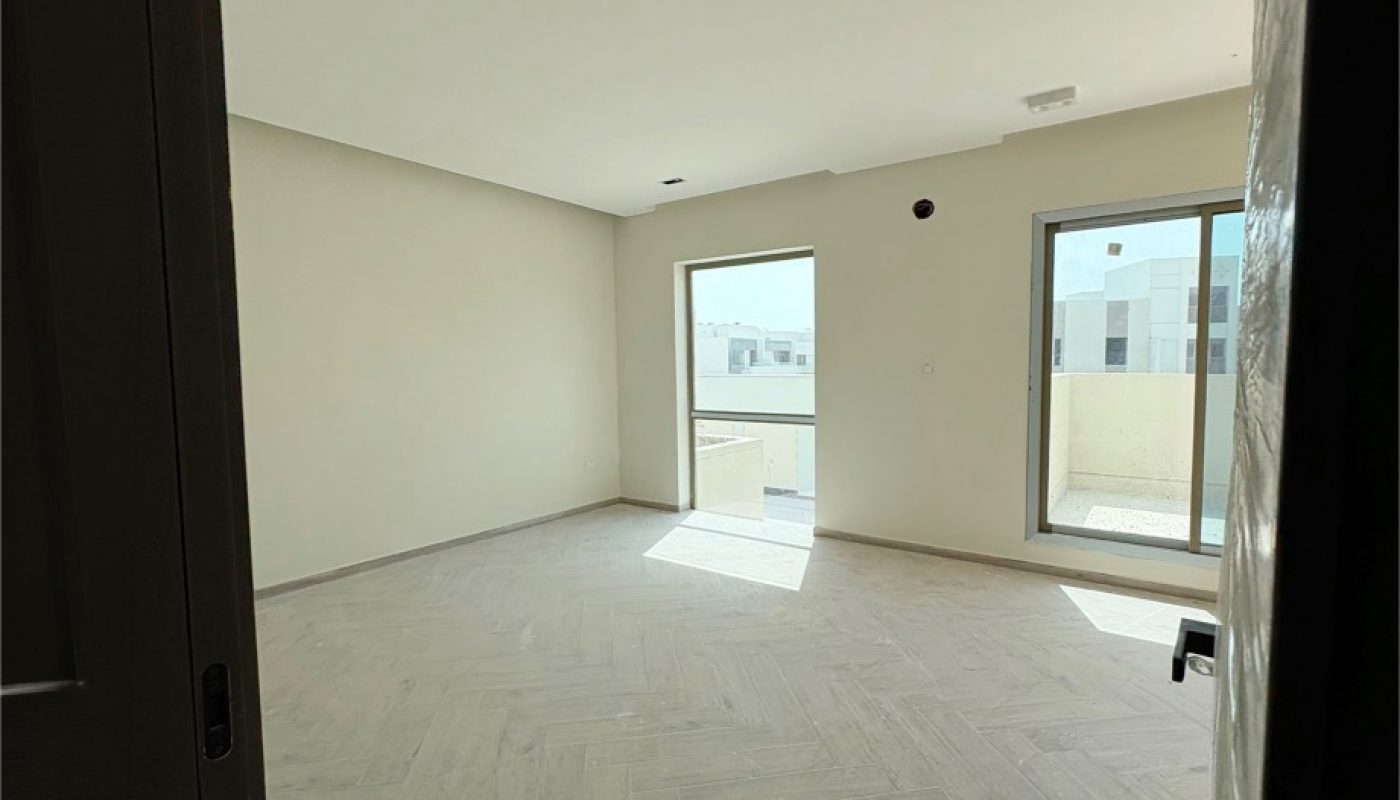 Empty room with light-colored walls, wood-patterned flooring, two large windows, and a sliding glass door opening to a balcony. Natural daylight fills the space.