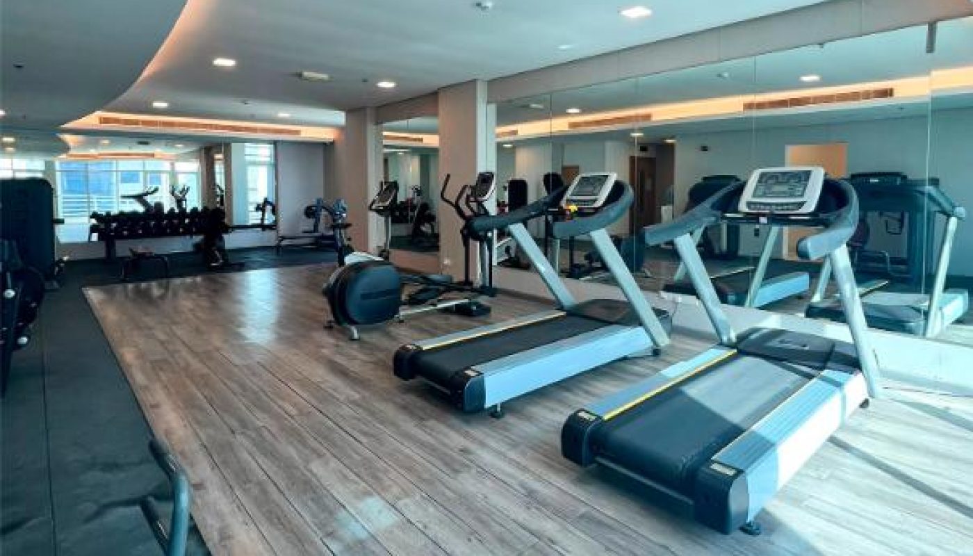 Modern gym room with treadmills, elliptical machines, weight benches, and large mirrors. Natural sunlight enters through floor-to-ceiling windows on one side.
