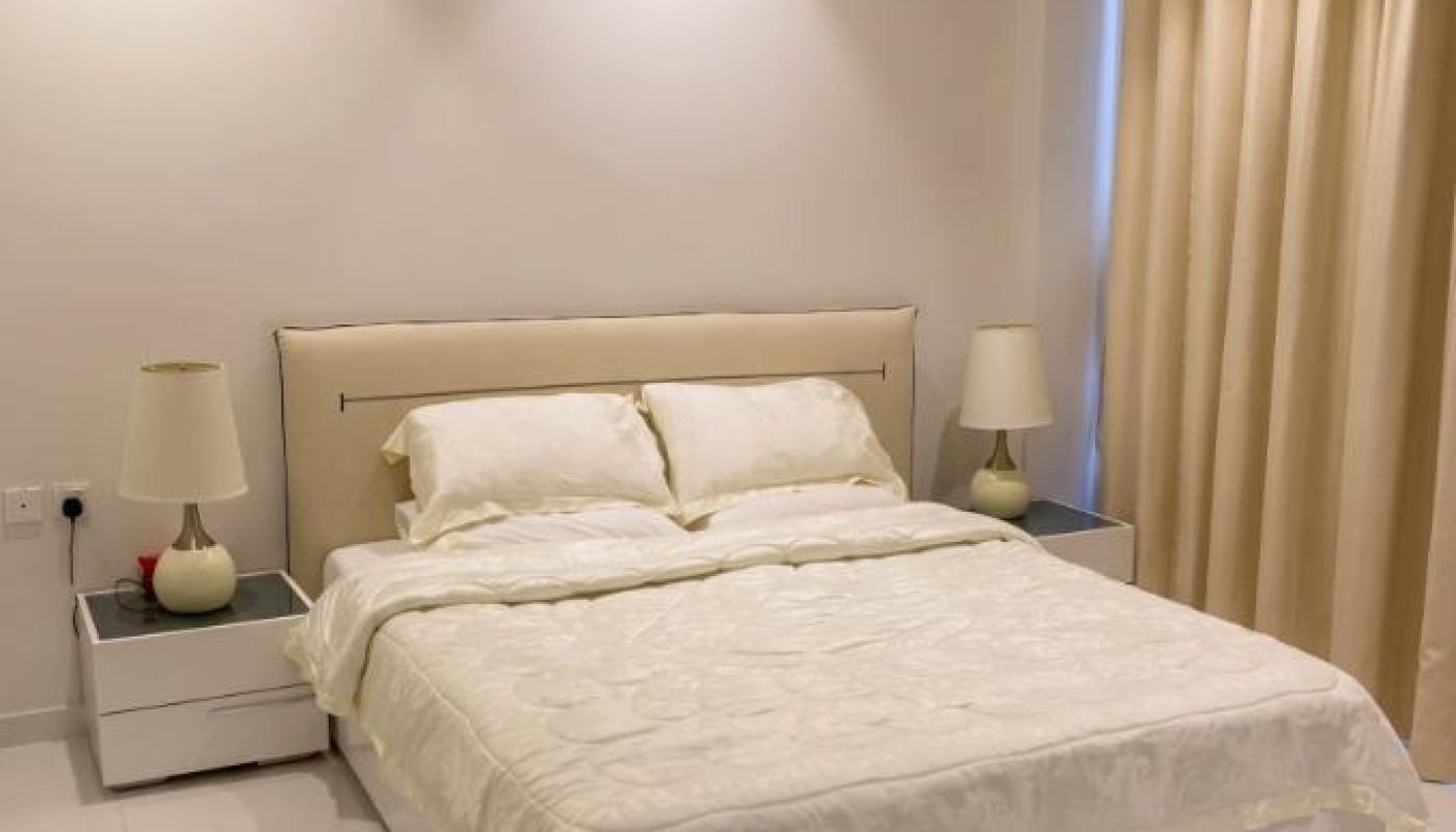 A neatly made bed with white bedding is centered between two nightstands with lamps in a minimalist bedroom with cream-colored curtains and tile flooring.