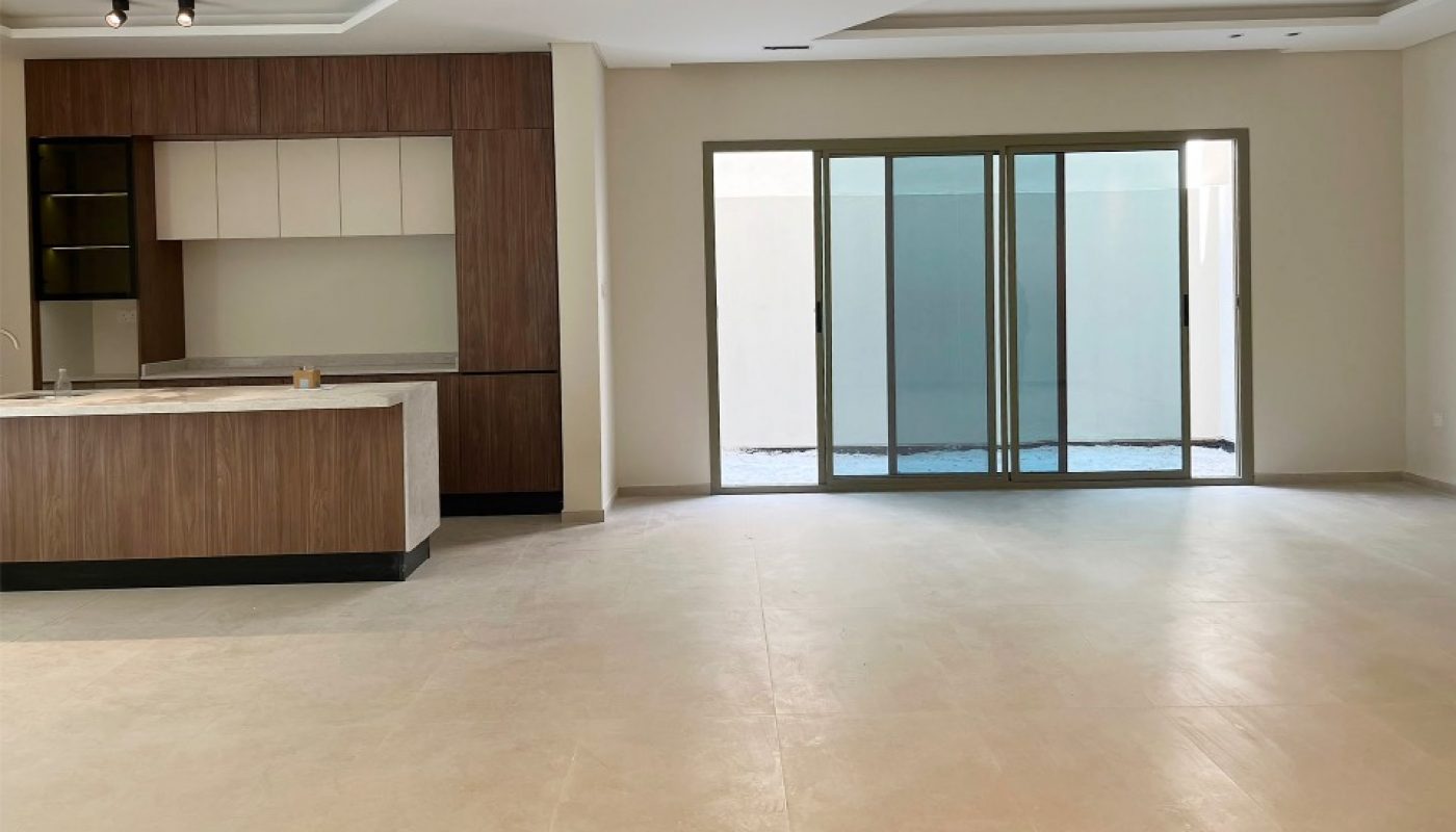 Spacious, unfurnished open-plan room with light floors, a modern kitchen with wood cabinets, and large glass sliding doors leading to an outdoor area.