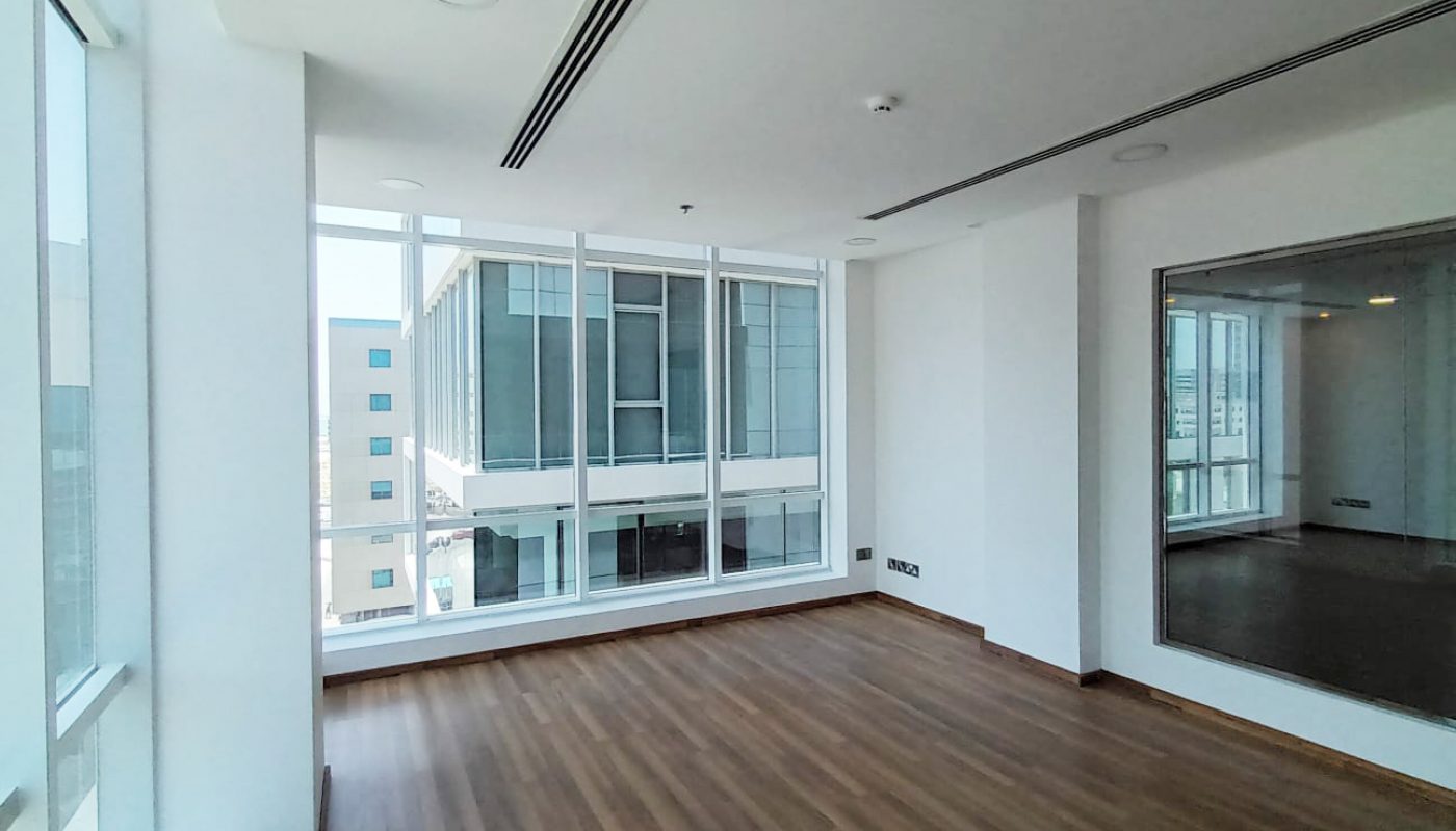 Empty room with wooden floor, large windows showing adjacent building, white walls, and a wall-mounted mirror.