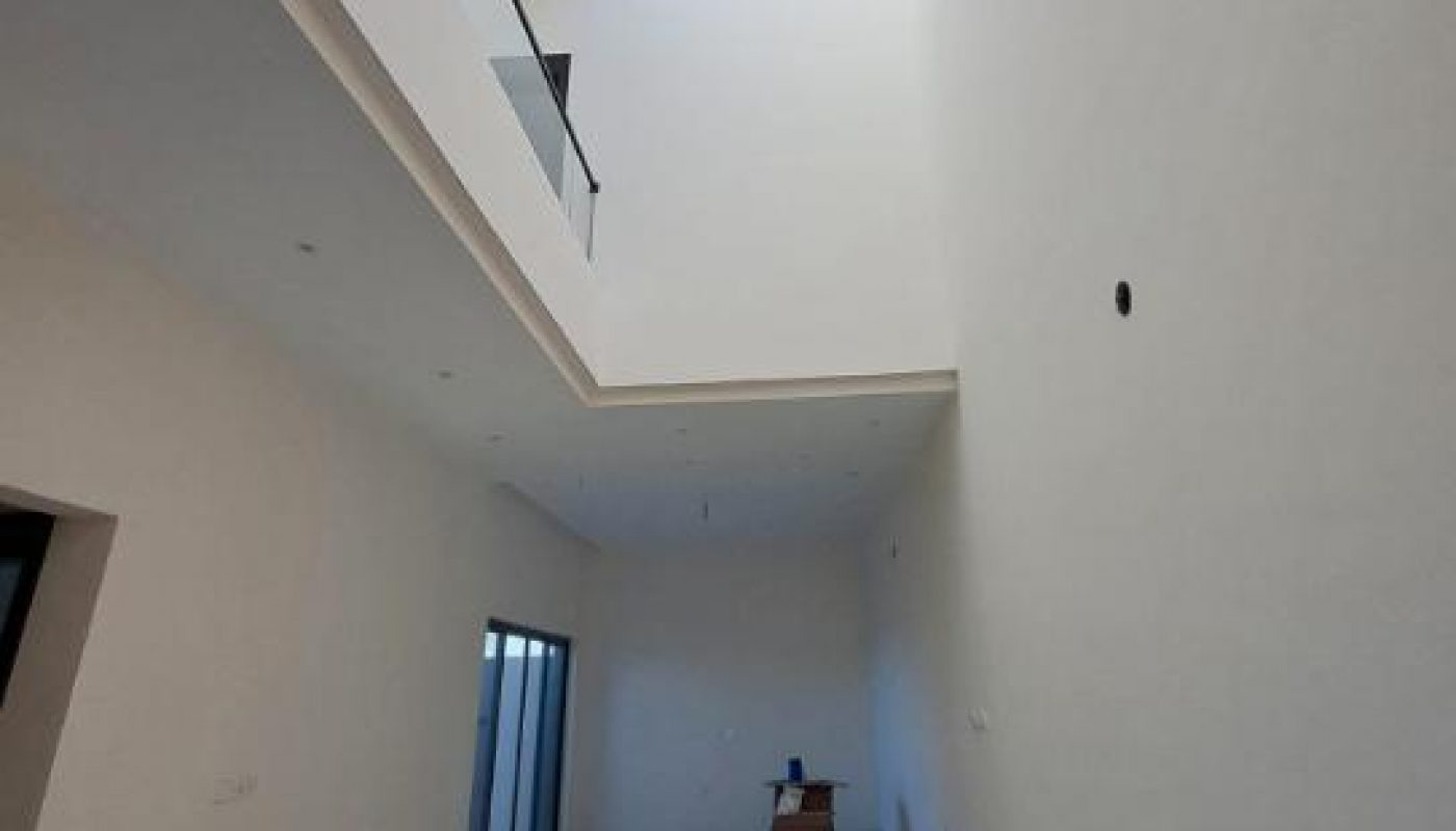 A tall, white-walled room with a skylight ceiling, glass balcony railing above, sliding glass doors on the left, and a small pile of items on the floor at the far end.