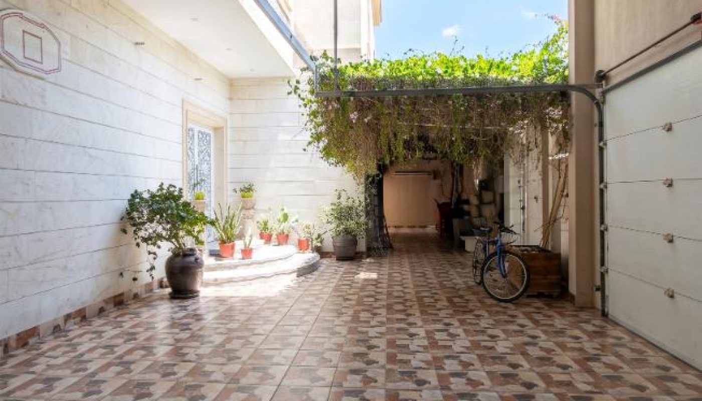 Open garage with tiled floor, potted plants along the left wall, a bicycle, and a leafy vine canopy at the far end.