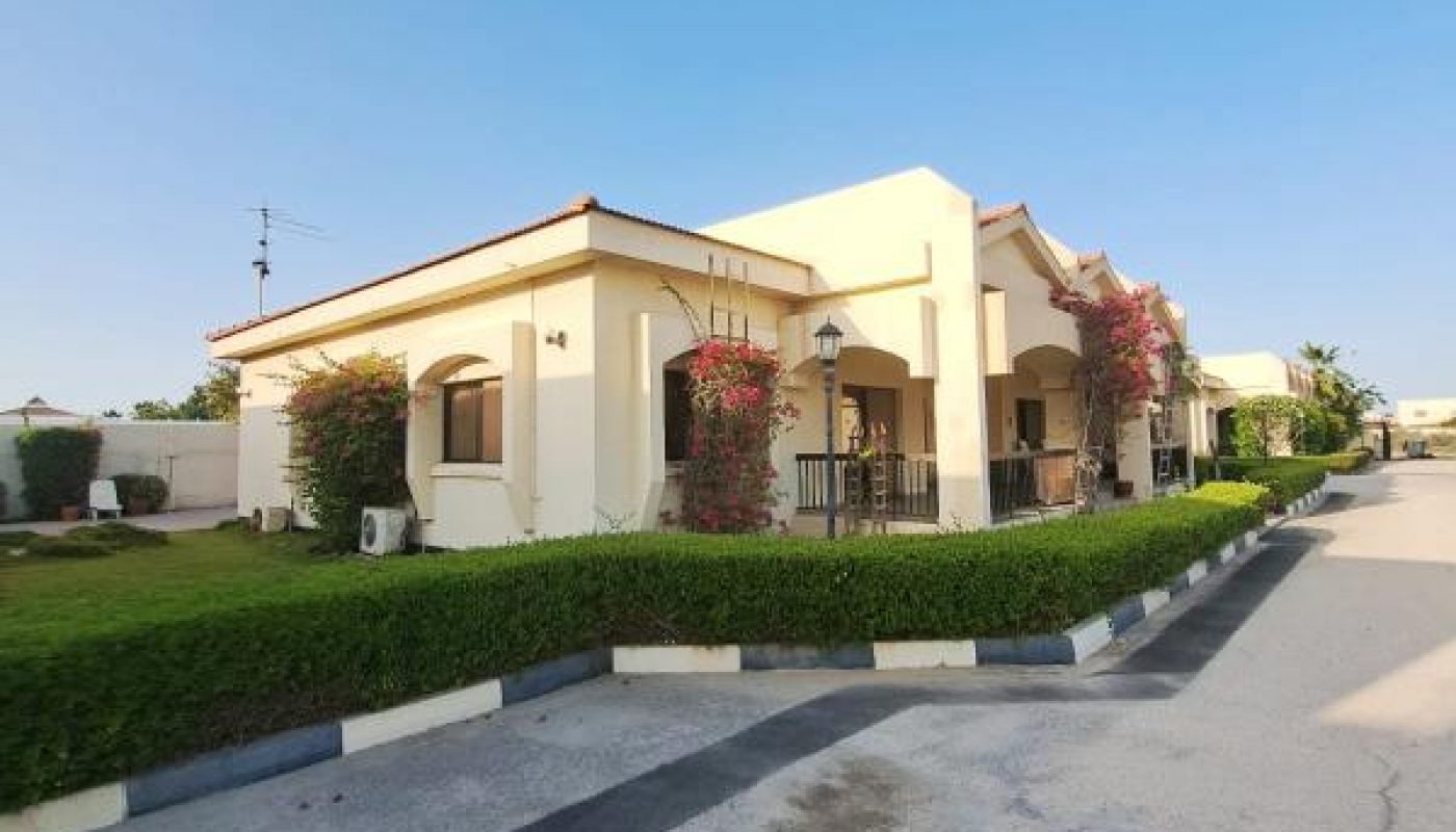 Single-story cream-colored house with a red-tiled roof, arched doorways, trimmed hedges, flowering bushes, and a paved driveway under a clear blue sky.