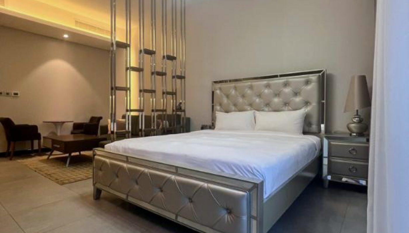 A modern bedroom with a metallic upholstered bed, matching nightstands, pendant lights, and a small dining area separated by vertical metal rods.