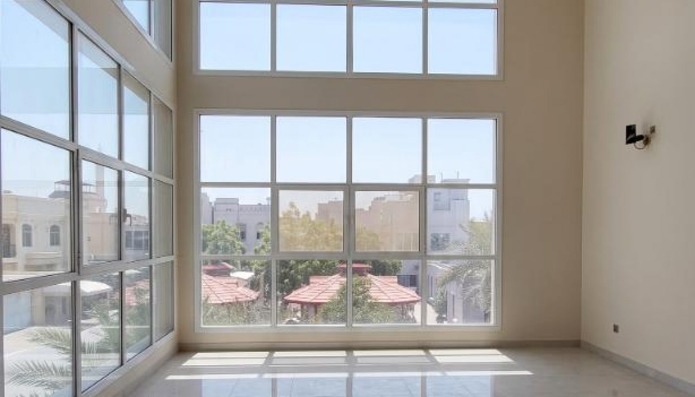 A spacious, empty room with large floor-to-ceiling windows, white tile flooring, beige walls, and an exterior view of buildings and trees.
