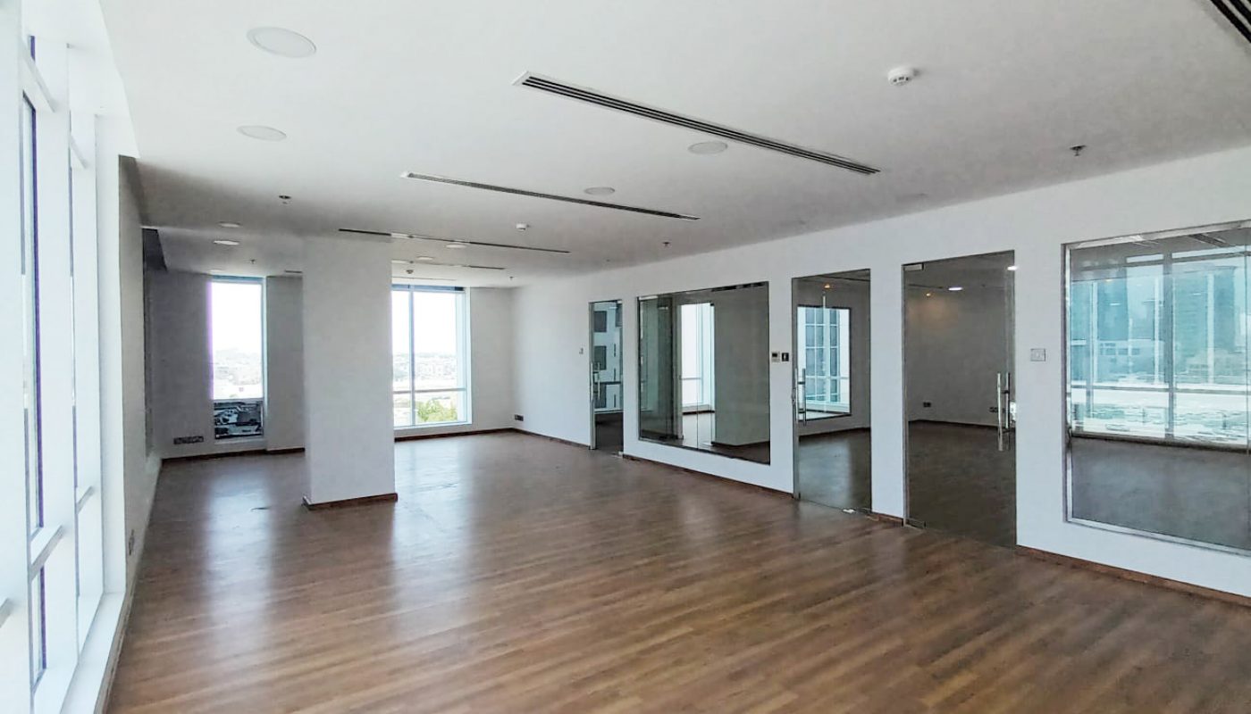 Empty room with large windows, wooden flooring, white walls, ceiling lights, and mirrored panels reflecting the space and exterior buildings.