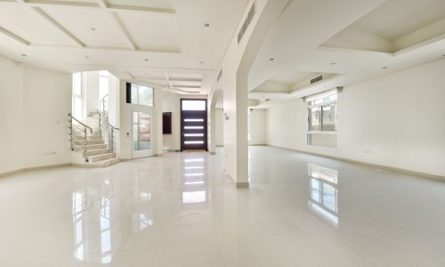 Spacious, brightly lit semi-furnished room with shiny white tiled floors, white walls, and a staircase at the back.