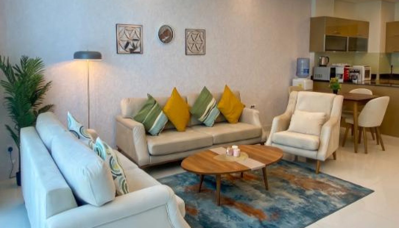 Modern living room with beige sofas, colorful cushions, a blue rug, coffee table, and an open kitchen in the background.