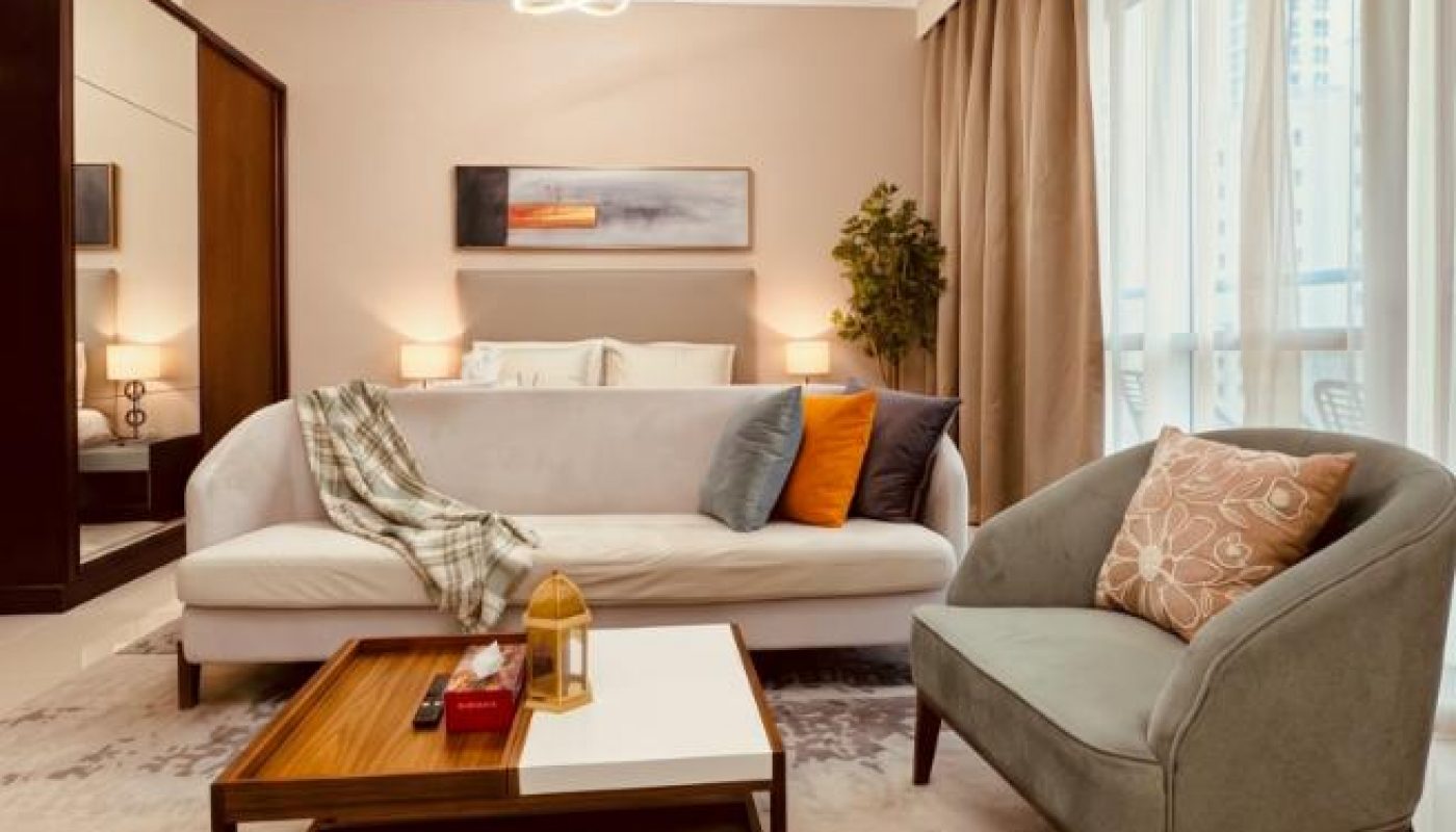 Modern hotel room with a bed, white sofa, grey armchair, wooden coffee table, neutral decor, large windows with sheer curtains, and warm ambient lighting.