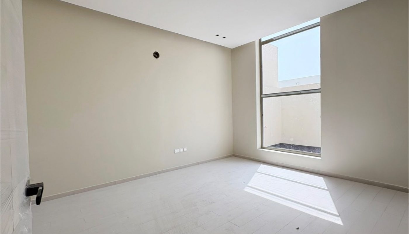 Empty room with beige walls, light-colored tiled floor, a large window letting in sunlight, and visible electrical outlets on the wall.