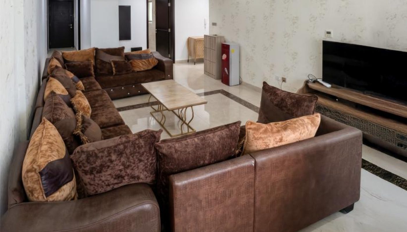 A modern living room with a large brown sectional sofa, a marble coffee table, a TV on a wooden stand, and glossy tiled flooring.