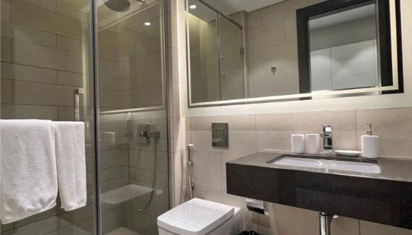 Modern bathroom with a glass shower enclosure, wall-mounted toilet, large mirror, sink with toiletries, towel rack, and a waste bin on a light-colored tiled floor.