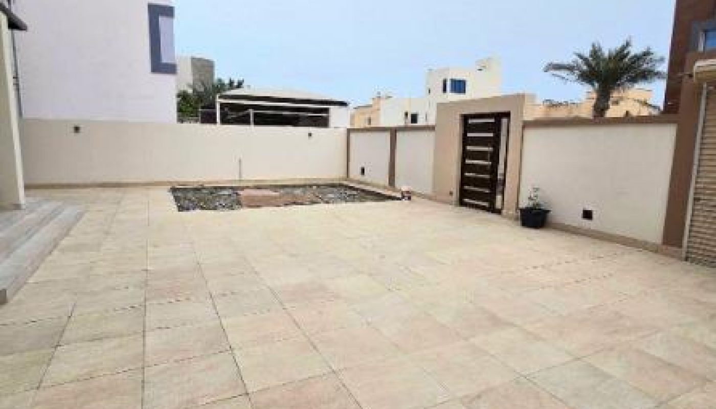 Spacious tiled patio with a central garden bed, surrounded by high walls and a modern gate, adjacent to white and brown buildings.