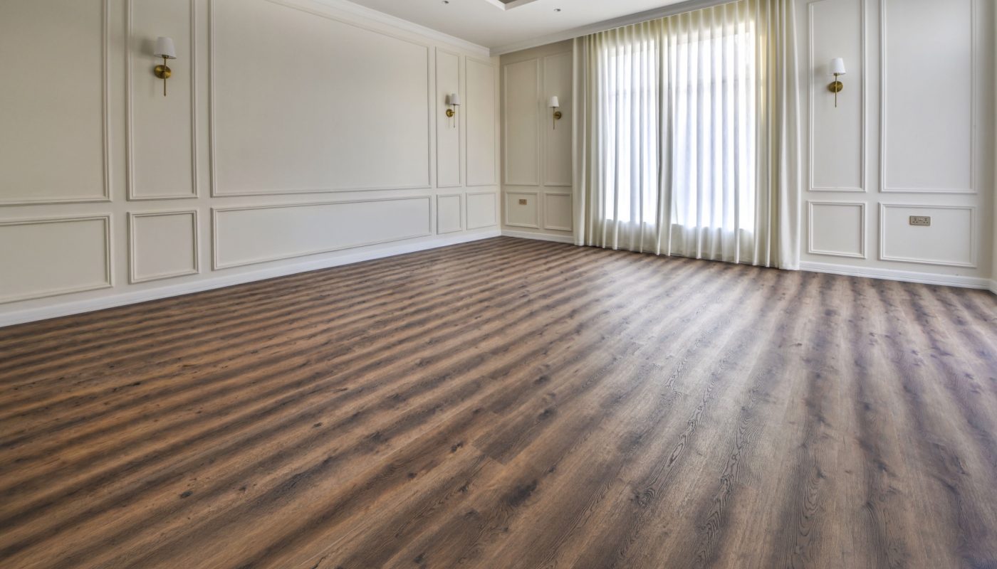 Spacious empty room with wood flooring, white paneled walls, ceiling lights, and large window covered by sheer white curtains.