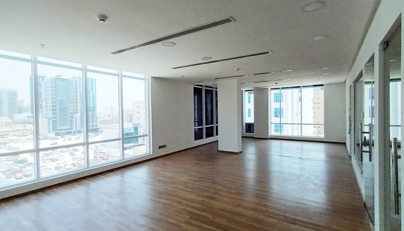Spacious empty office with large windows, wooden floors, and city buildings visible outside during daytime.