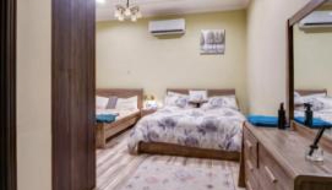 A neatly arranged bedroom with two beds, a wardrobe, a dresser with a mirror, an air conditioner, and a patterned rug on the wooden floor.