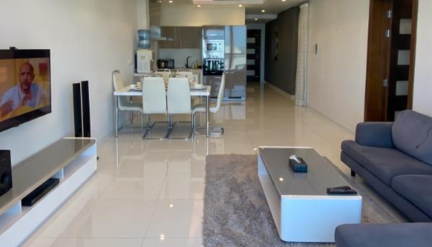 Modern open-plan living room and kitchen with white tile floors, gray sofa, TV on the wall, dining table, and stainless steel appliances.