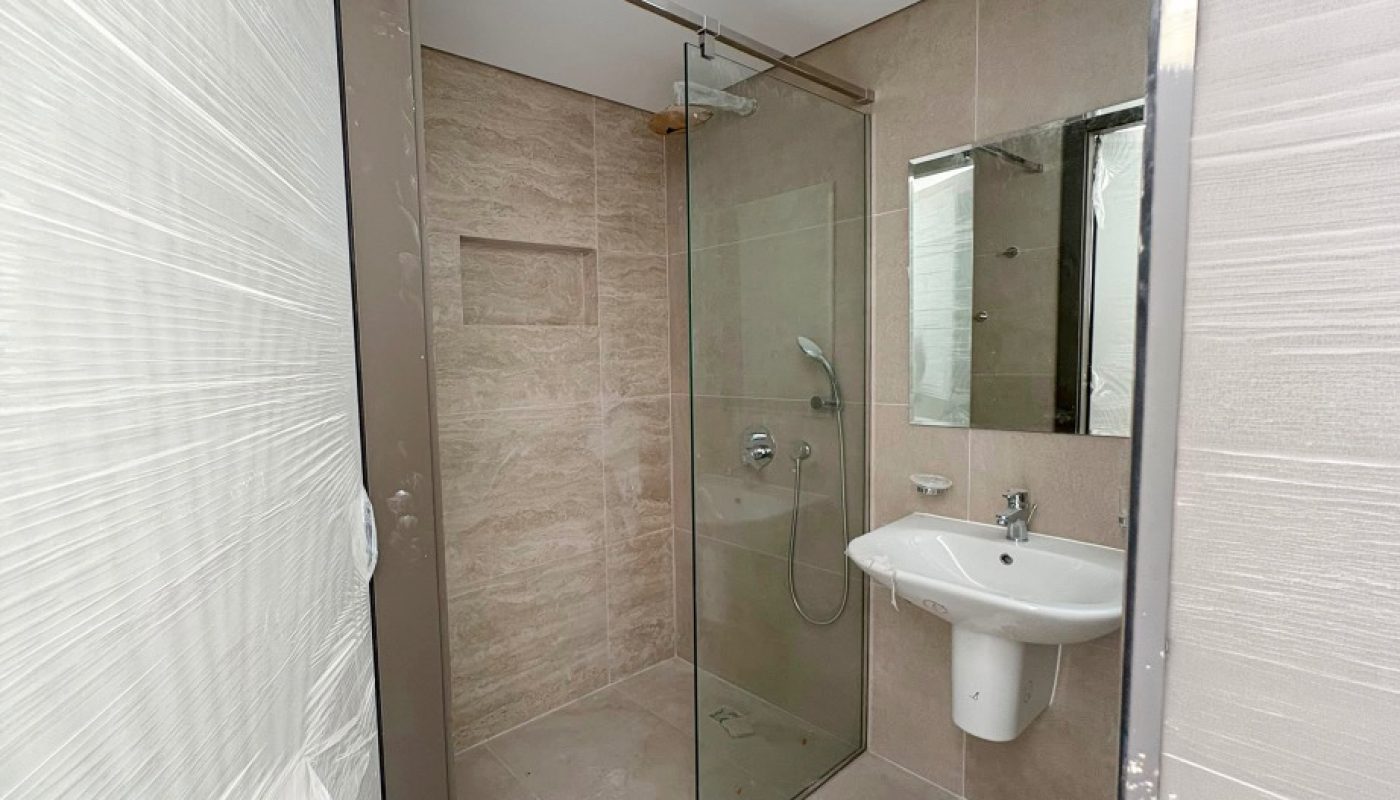 Modern bathroom with beige tiles, walk-in glass shower, wall-mounted sink, and mirror. Doorframe and trim appear to be under protective plastic covering.