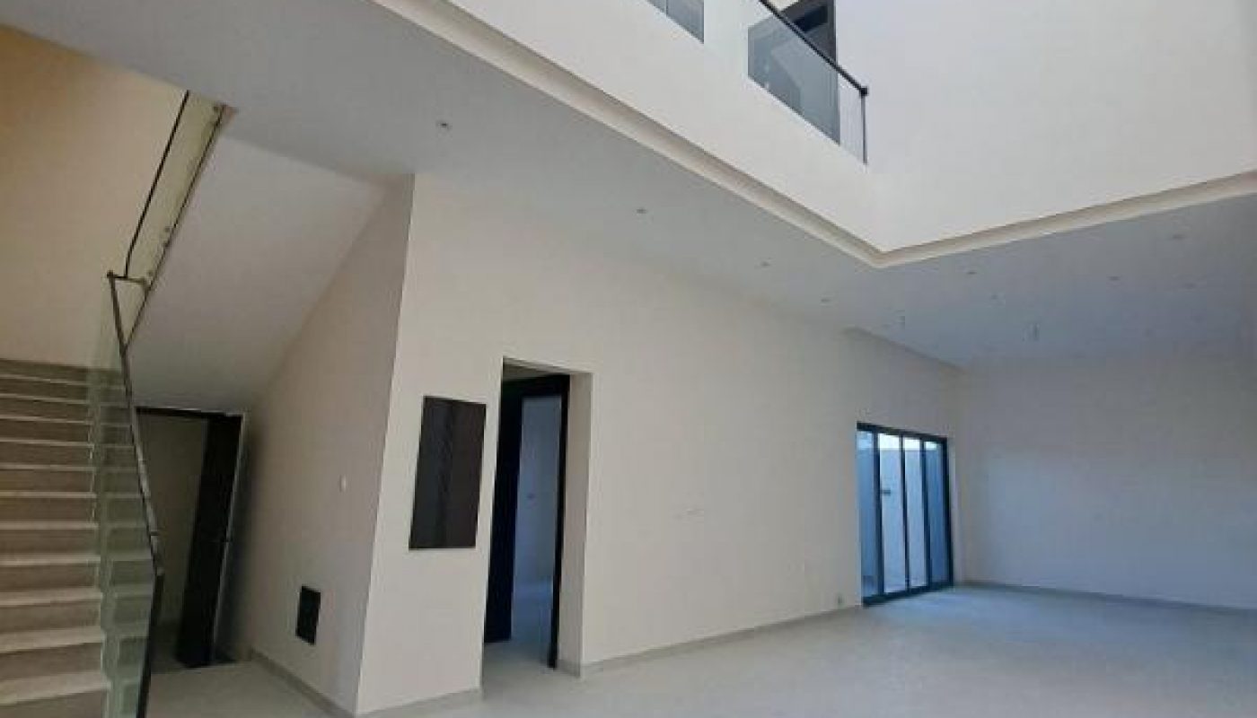 Spacious, modern interior with white walls, high ceiling, large windows, glass balcony railing, and staircase to an upper floor.