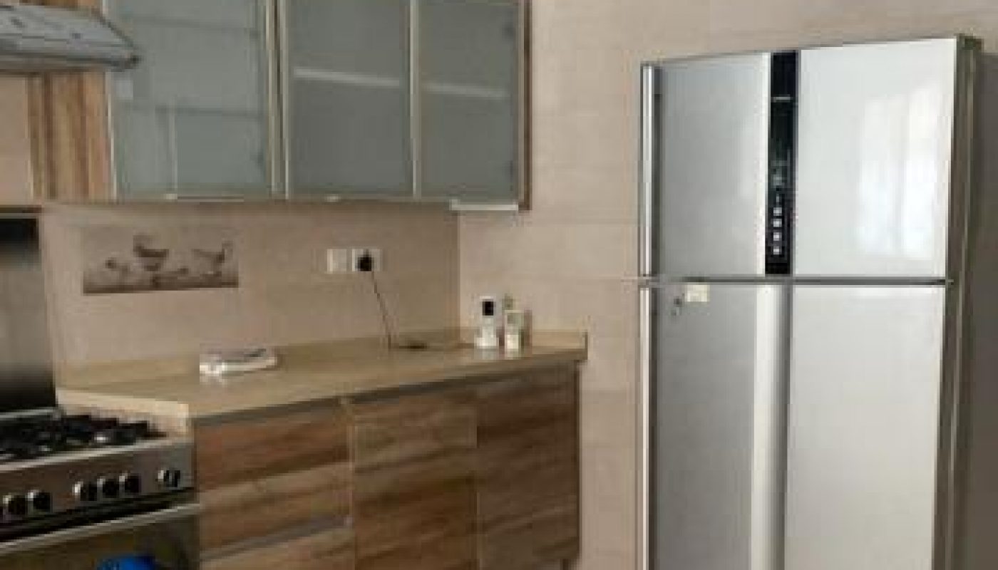 Modern kitchen with stainless steel refrigerator, oven, gas stove, and wooden cabinets against beige tiled walls and floor.