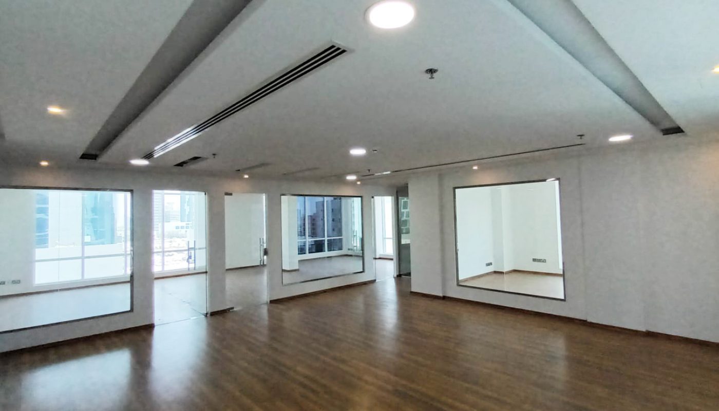 Spacious, empty room with wooden floors, large mirrors on the walls, and ceiling lights. Windows let in natural light from outside.