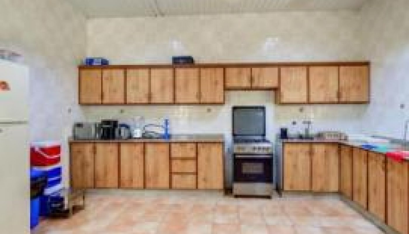 Spacious kitchen with tiled floor, wooden cabinets, stove, fridge, and various appliances lining the back wall under fluorescent lighting.