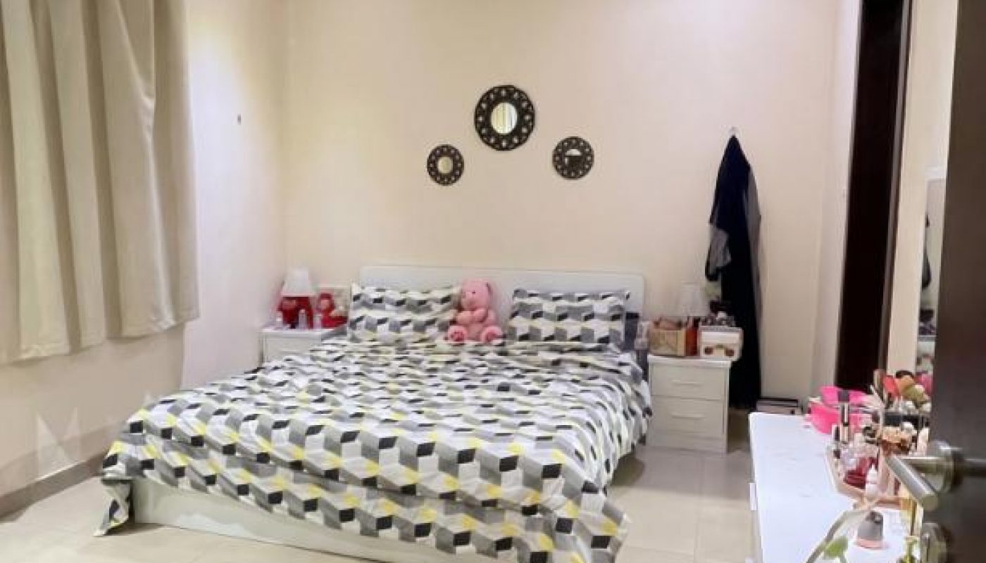 A neatly organized bedroom with a double bed, geometric-patterned bedding, nightstands, circular wall mirrors, and a dresser with various items. Light curtains cover the window.