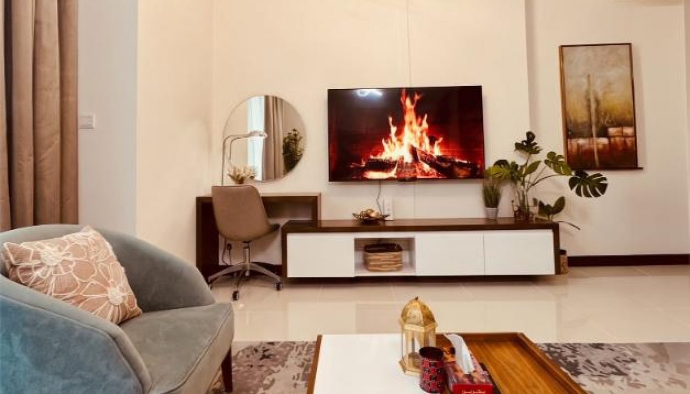 Modern living room with a blue armchair, coffee table, TV displaying a fireplace, desk with a mirror, houseplants, and contemporary lighting fixture.