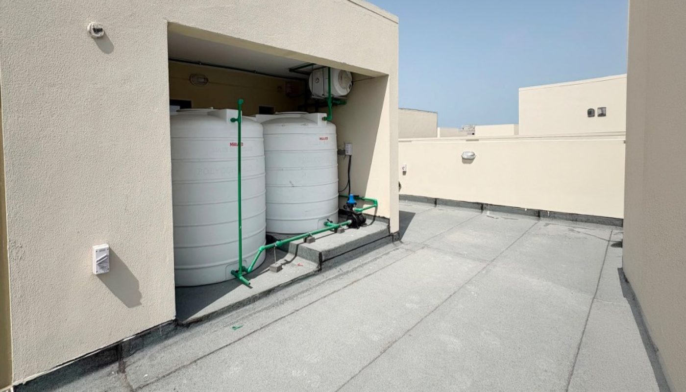 View of a flat rooftop with two large white water tanks and equipment stored in a recessed area against a beige wall under a clear blue sky.
