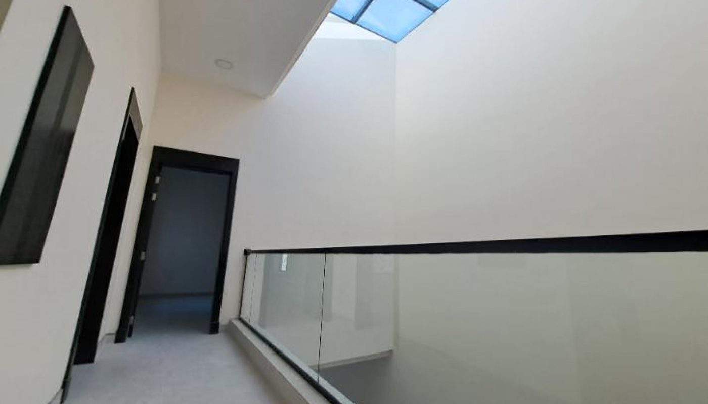 A modern hallway with a glass railing and a large skylight, leading to a dark doorway at the end.