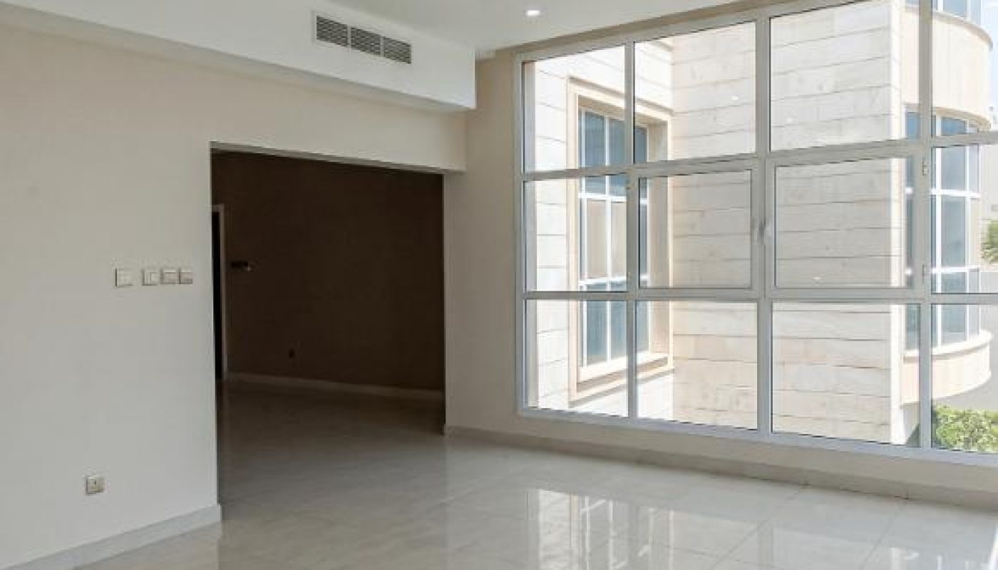 Empty room with large tile floor, ceiling light fixture, white walls, and a wall of tall windows showing part of a building exterior.
