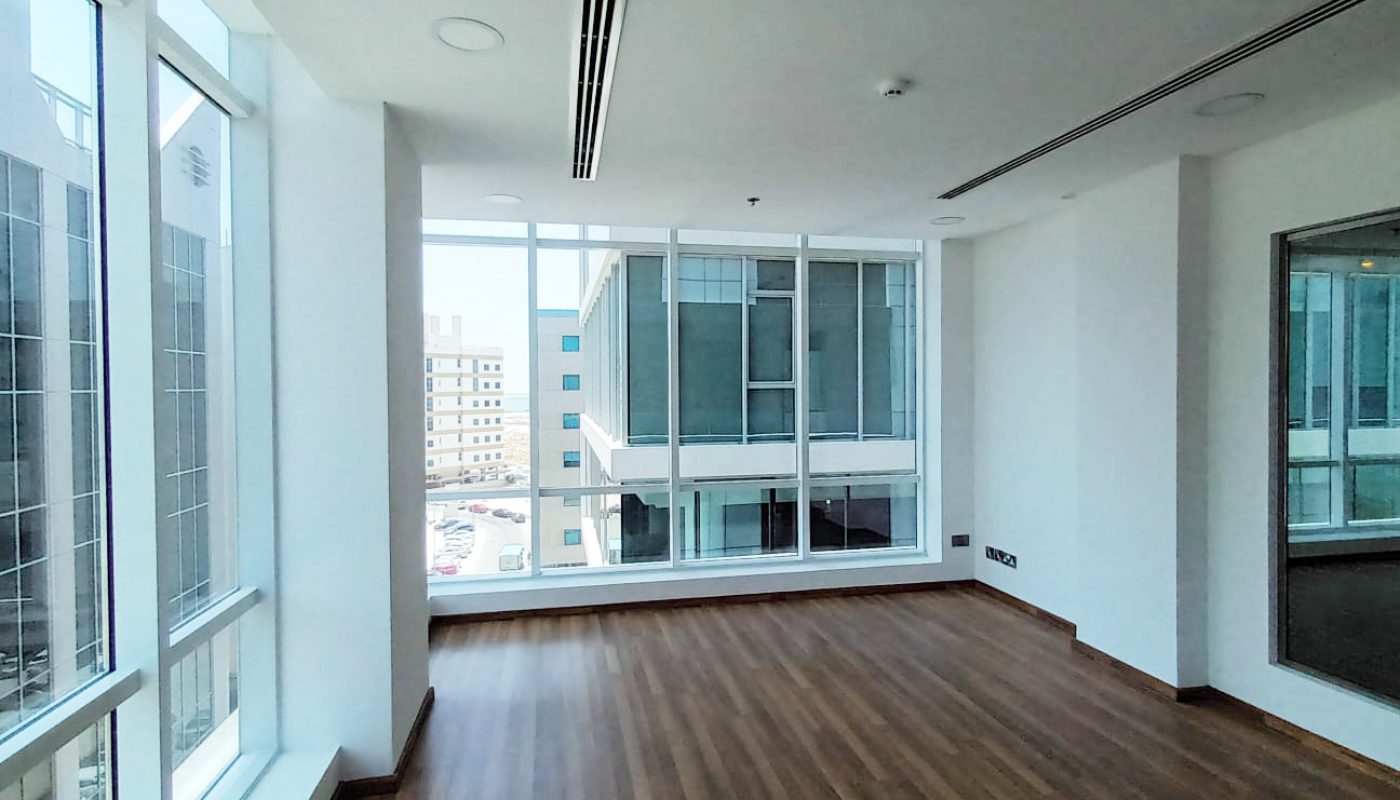 Empty office room with large floor-to-ceiling windows, wood flooring, white walls, and a view of nearby modern buildings outside.