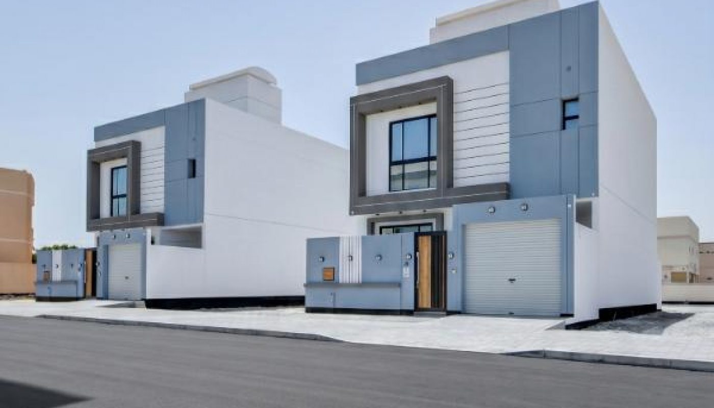 Two modern, white and blue, two-story houses with garages, line a paved street under a clear blue sky.