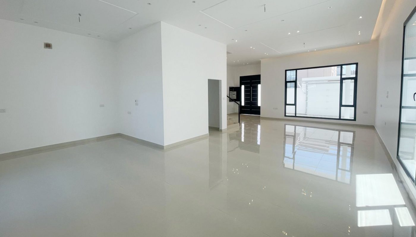 Spacious, empty room with white walls, glossy floor, large windows, and recessed lighting. Natural light enters through the windows and glass doors.