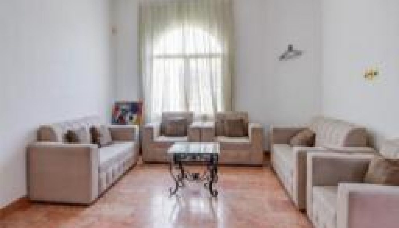 A living room with three beige sofas, a glass coffee table, a window with sheer curtains, and marble tile flooring.