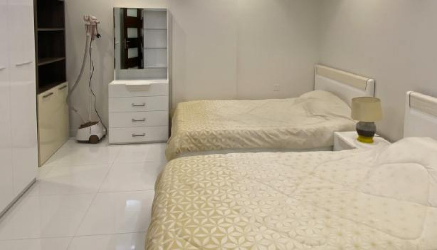A clean, modern bedroom with two single beds, a white dresser with a mirror, a nightstand with a lamp, and tiled floors under recessed ceiling lights.