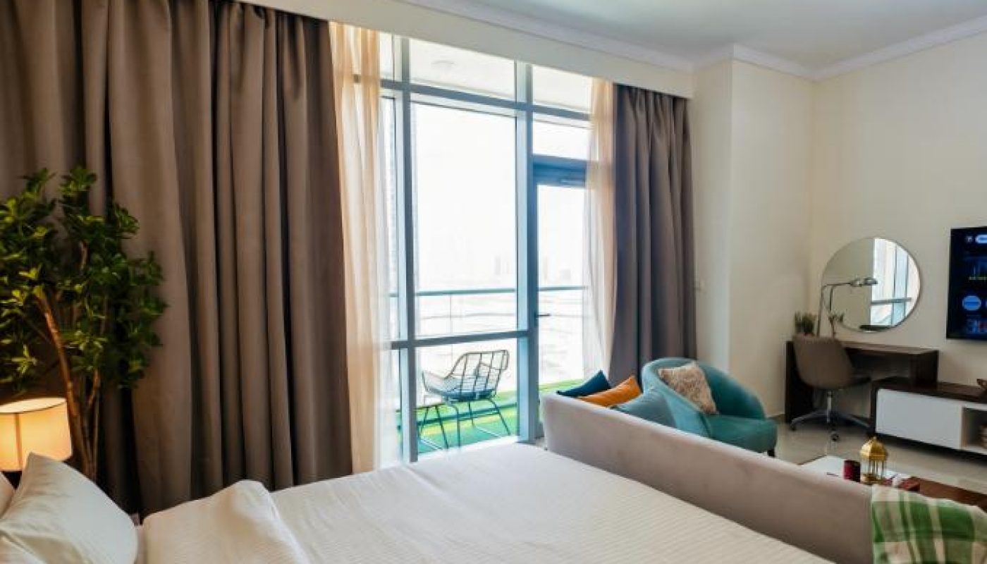 Modern bedroom with a double bed, green chair, vanity desk, TV, potted plant, large window, and balcony. Neutral colors and contemporary lighting fixture are featured.