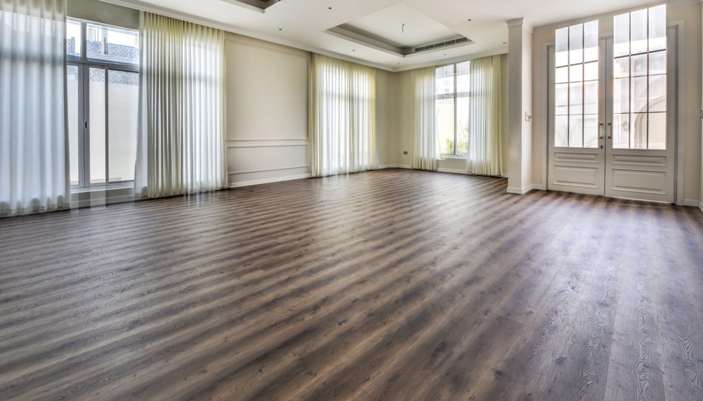 Large empty room with wood flooring, white walls, floor-to-ceiling windows, sheer curtains, and recessed ceiling features. Natural light fills the space.