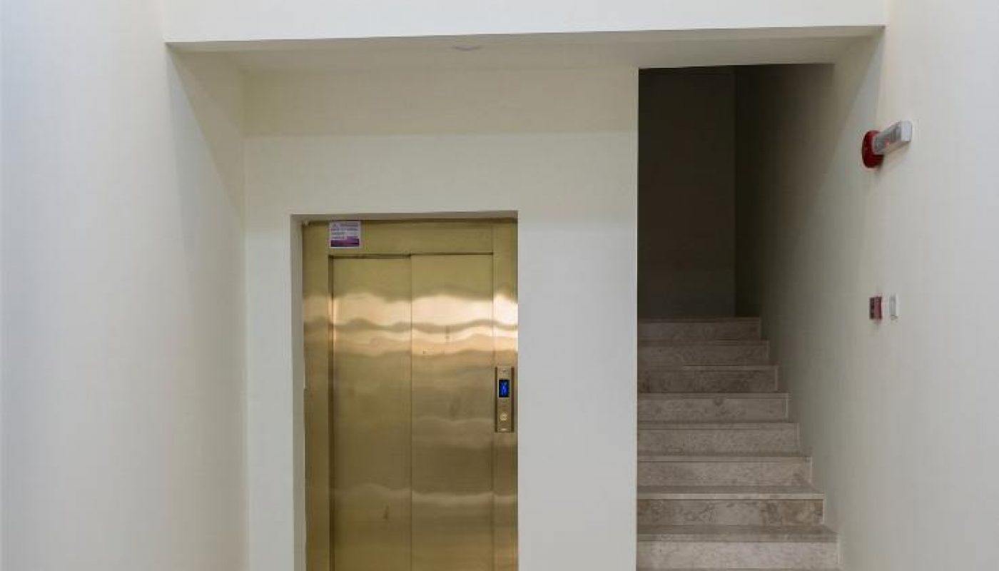 A hallway with a closed gold elevator door on the left and a staircase with light-colored steps on the right; tiled floor and white walls.