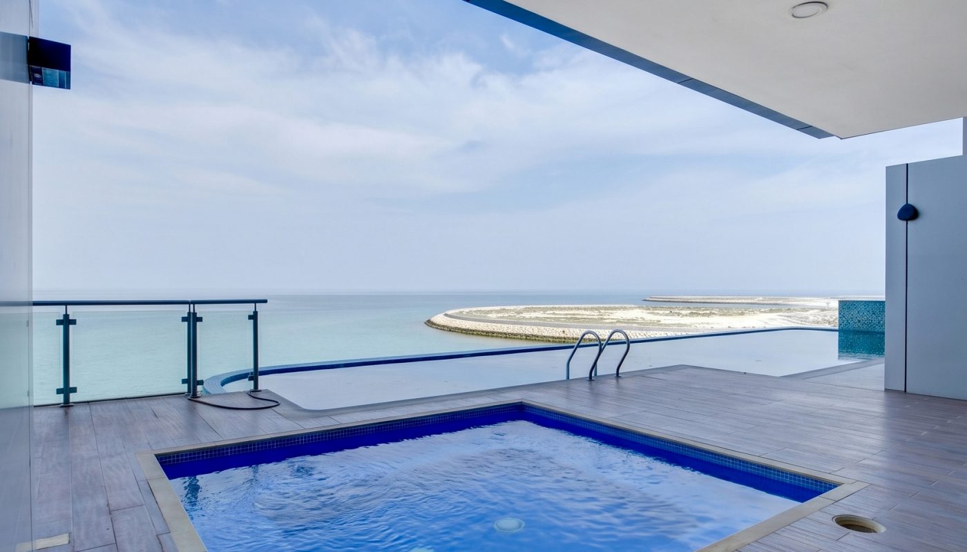 A rectangular swimming pool on a tiled deck overlooks the ocean, with a glass railing and a clear blue sky in the background.