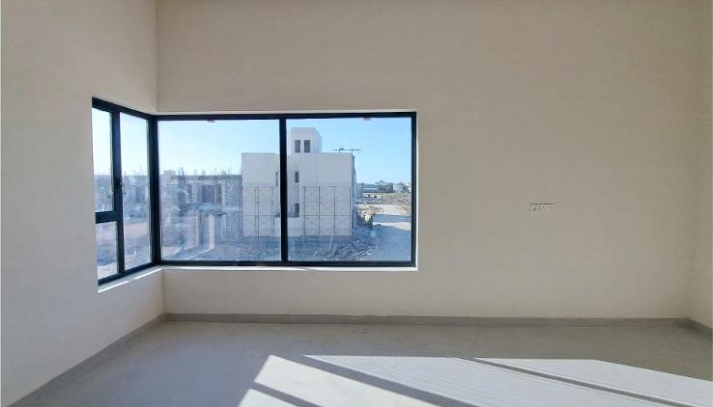 Empty room with beige walls, a large window with black frames, and sunlight casting shadows on the bare floor. A building is visible outside the window.
