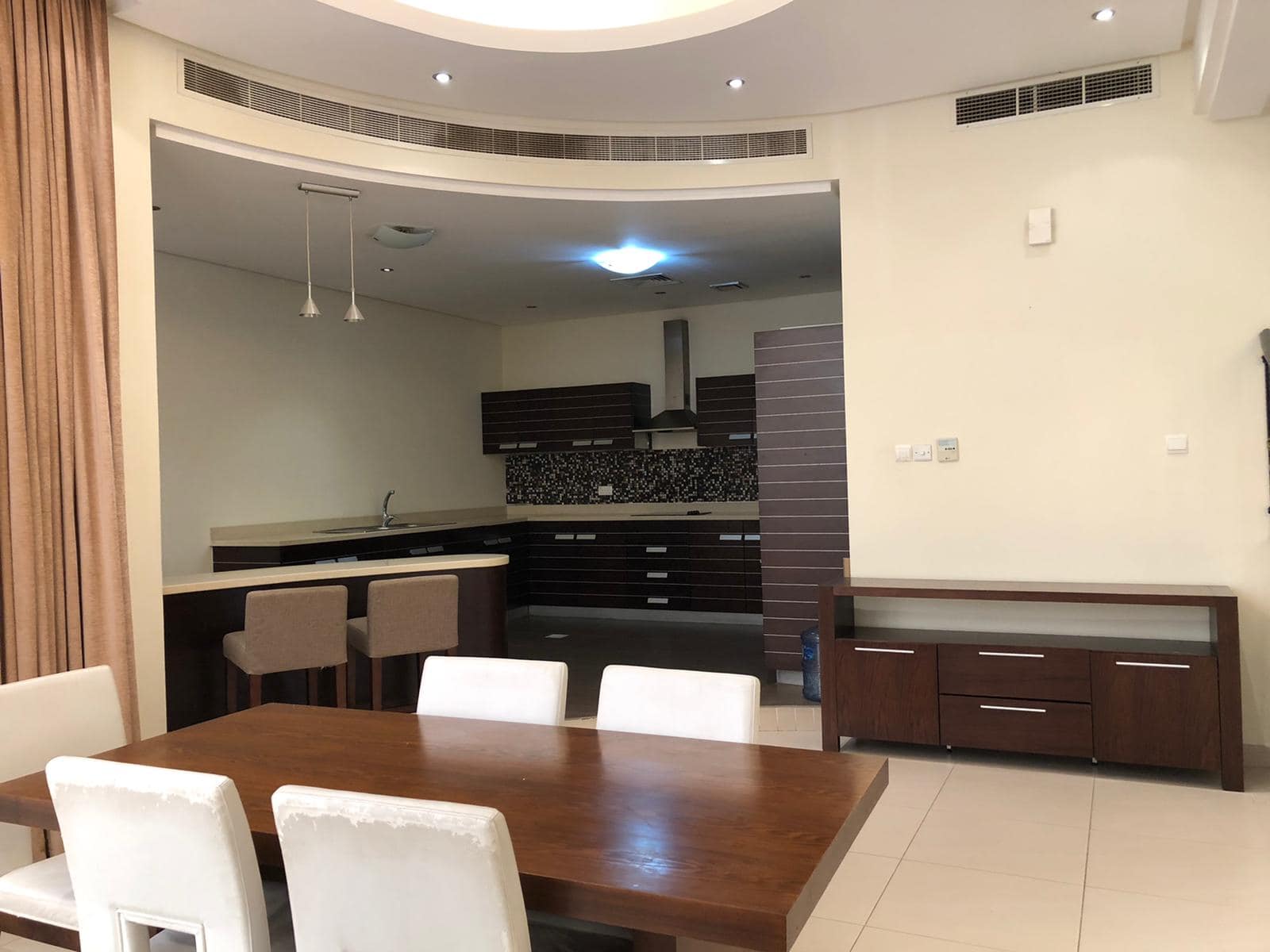 A modern dining area with a wooden table and white chairs, adjacent to a kitchen with dark cabinets and a breakfast bar. The ceiling has recessed lighting, and there are beige curtains on one side.