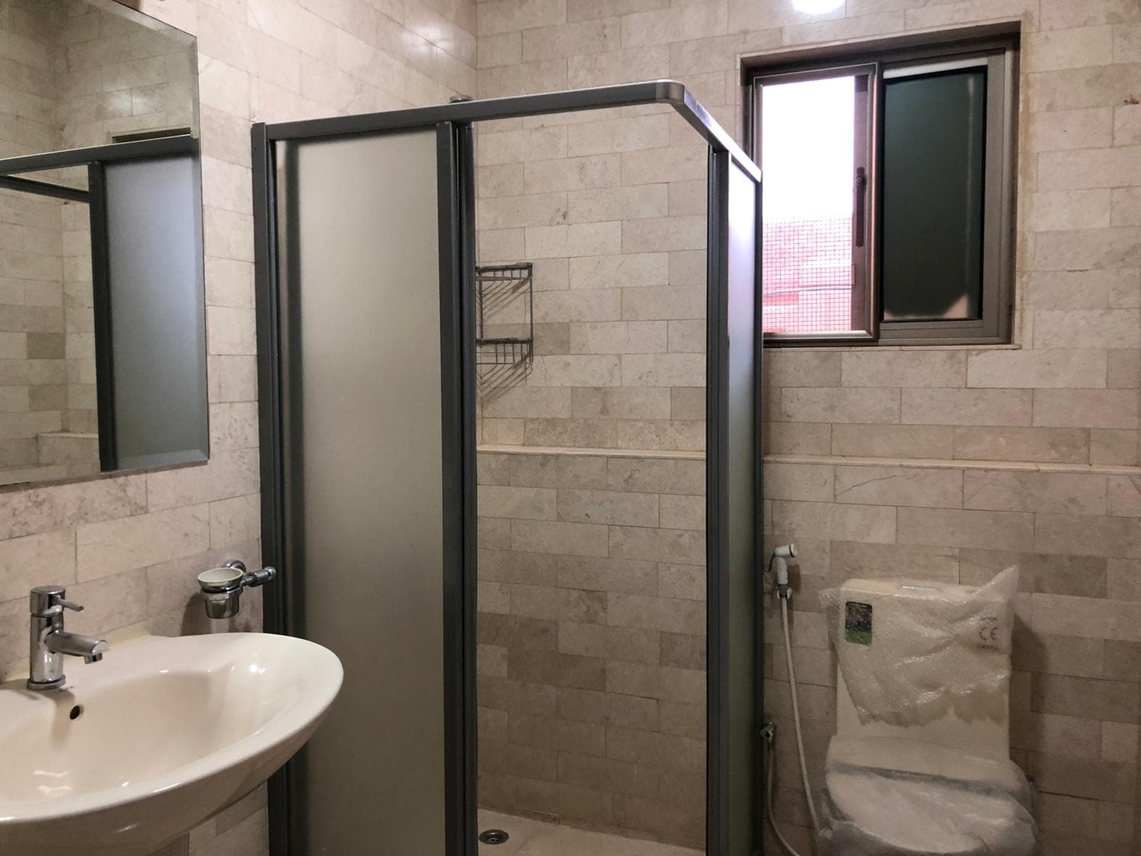 A bathroom with beige tiled walls, a walk-in shower with a glass door, a white sink with a wall-mounted faucet, a toilet, a small shelf, and a window with frosted glass.