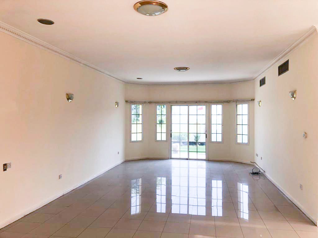 A large empty room with white walls, tiled floor, ceiling lights, and a set of French doors leading to a garden.