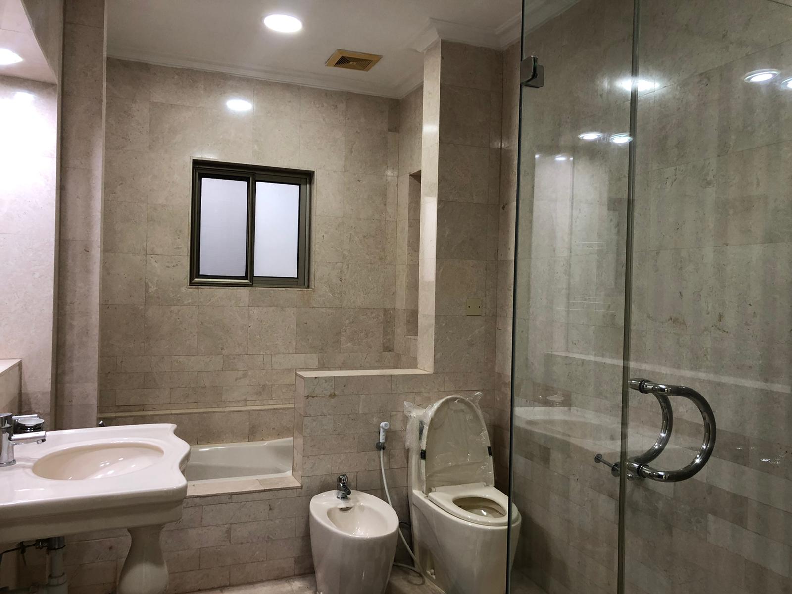 A bathroom with a sink, bathtub, toilet, bidet, and glass-enclosed shower. The walls and floor are tiled in beige. The room has recessed ceiling lights and a small window.