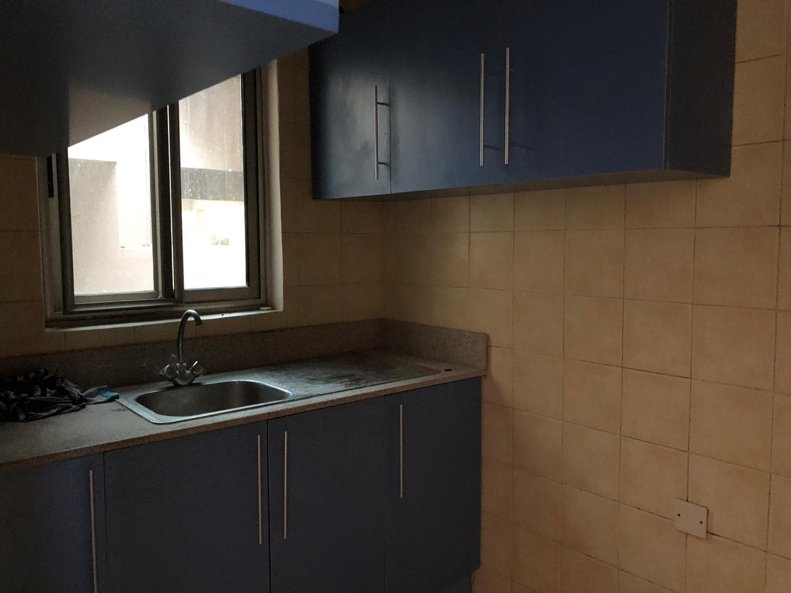 A small kitchen with beige tiled walls, blue cabinets, a stainless steel sink, and a window on the left.