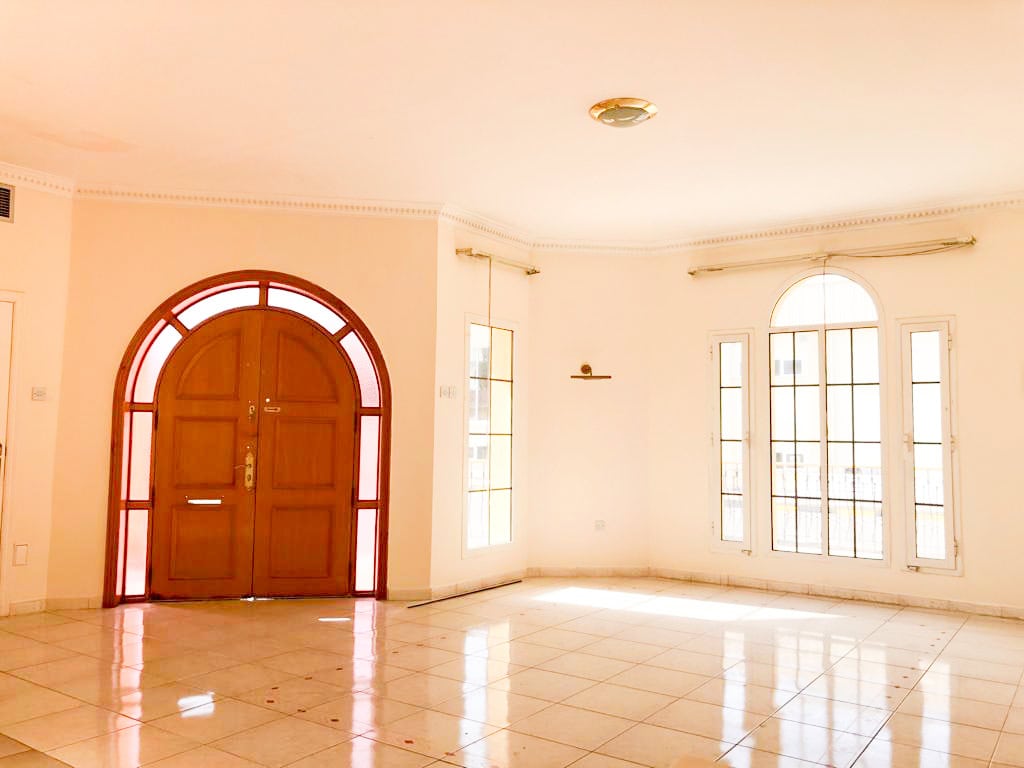 A spacious empty room with white tiled floor, large wooden front door, and tall arched windows allowing natural light to enter.